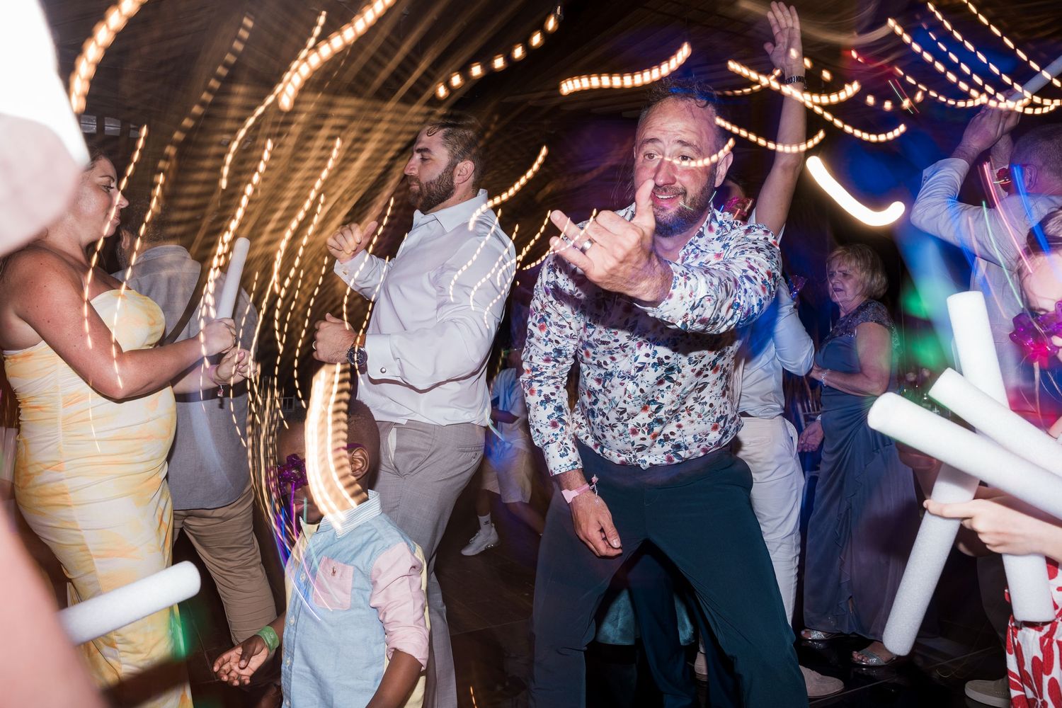 Man in floral shirt dances joyfully at a lively party surrounded by guests with glow sticks.