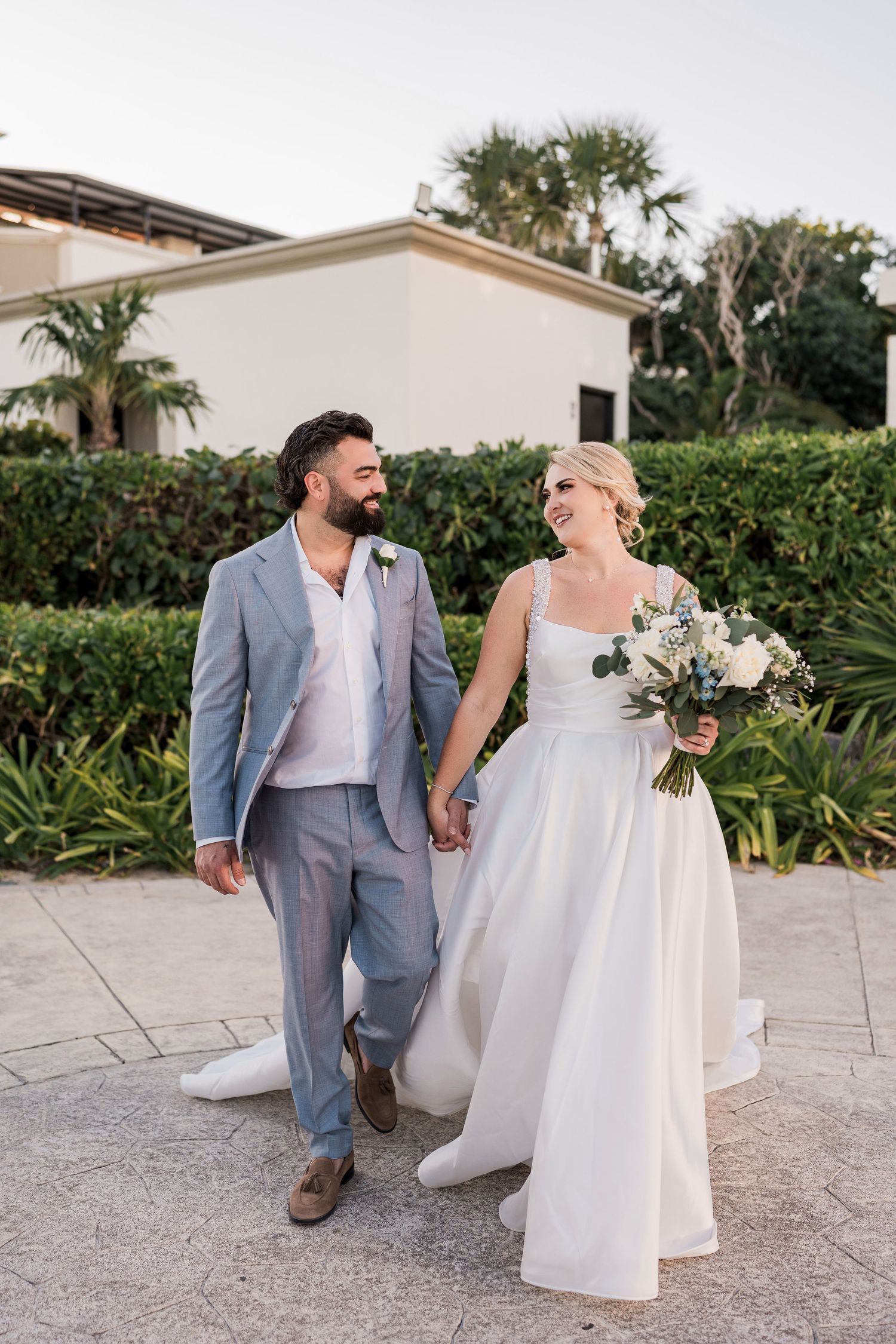 Bride and groom walk hand in hand outdoors, she holds white bouquet in wedding dress, he wears light blue suit.