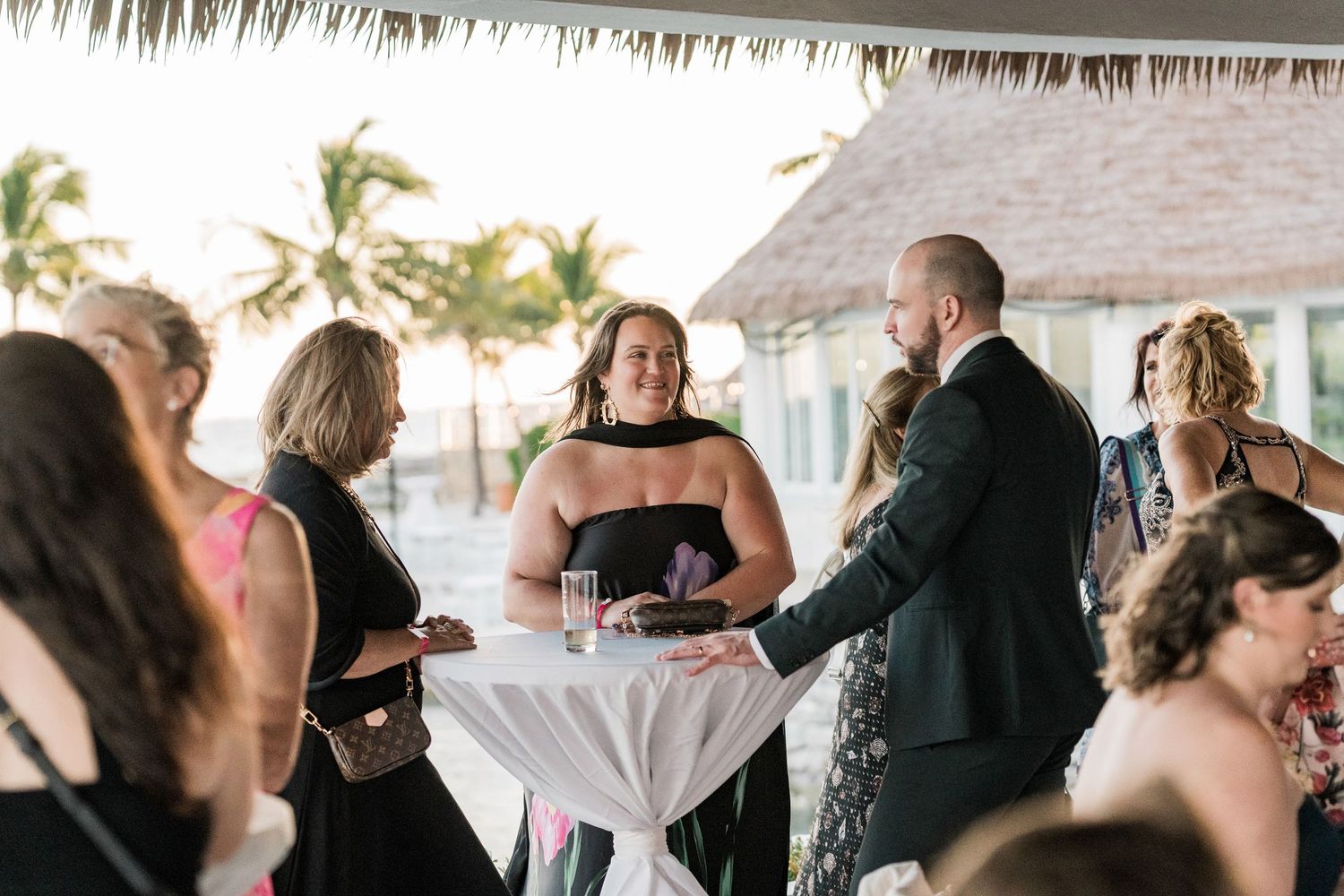 Guests mingle at an outdoor beachside wedding reception under a thatched roof, with palm trees visible in the background.