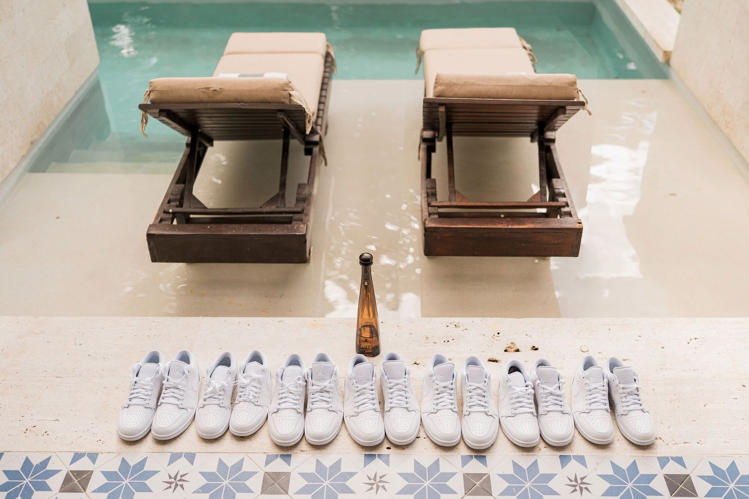 White sneakers displayed on a mosaic tile floor near a pool with wooden lounge chairs and a bottle.