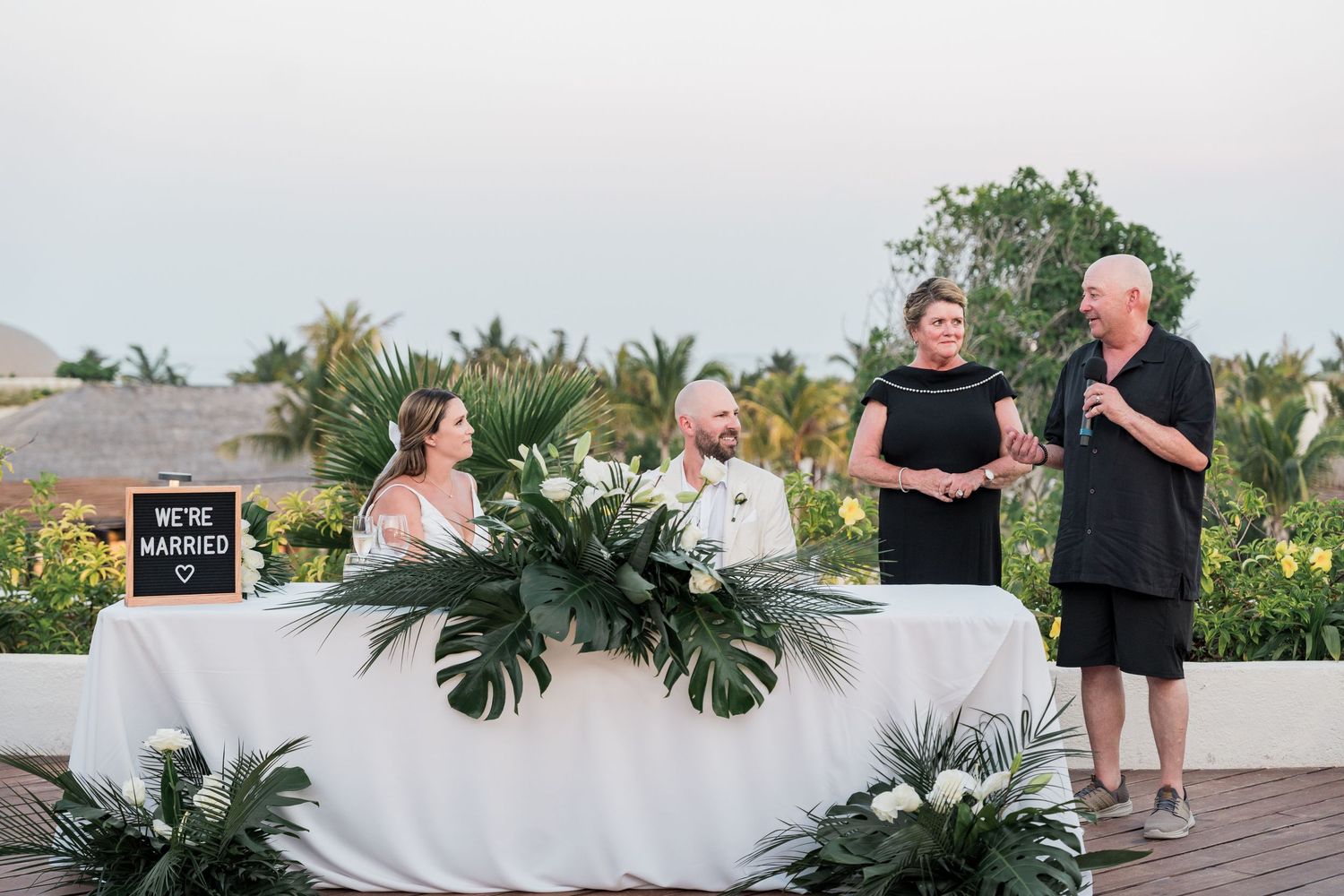 Outdoor wedding ceremony with couple at decorated table, officiant, and tropical greenery backdrop.