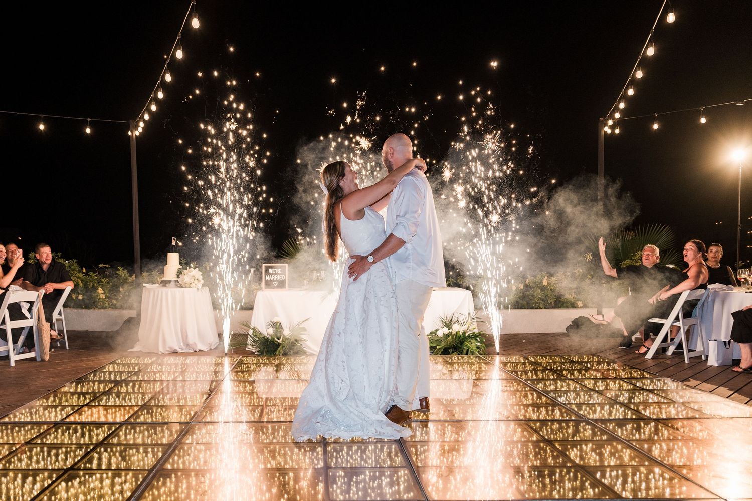 Couple shares first dance on illuminated floor with sparkling fireworks and string lights at elegant outdoor wedding reception.