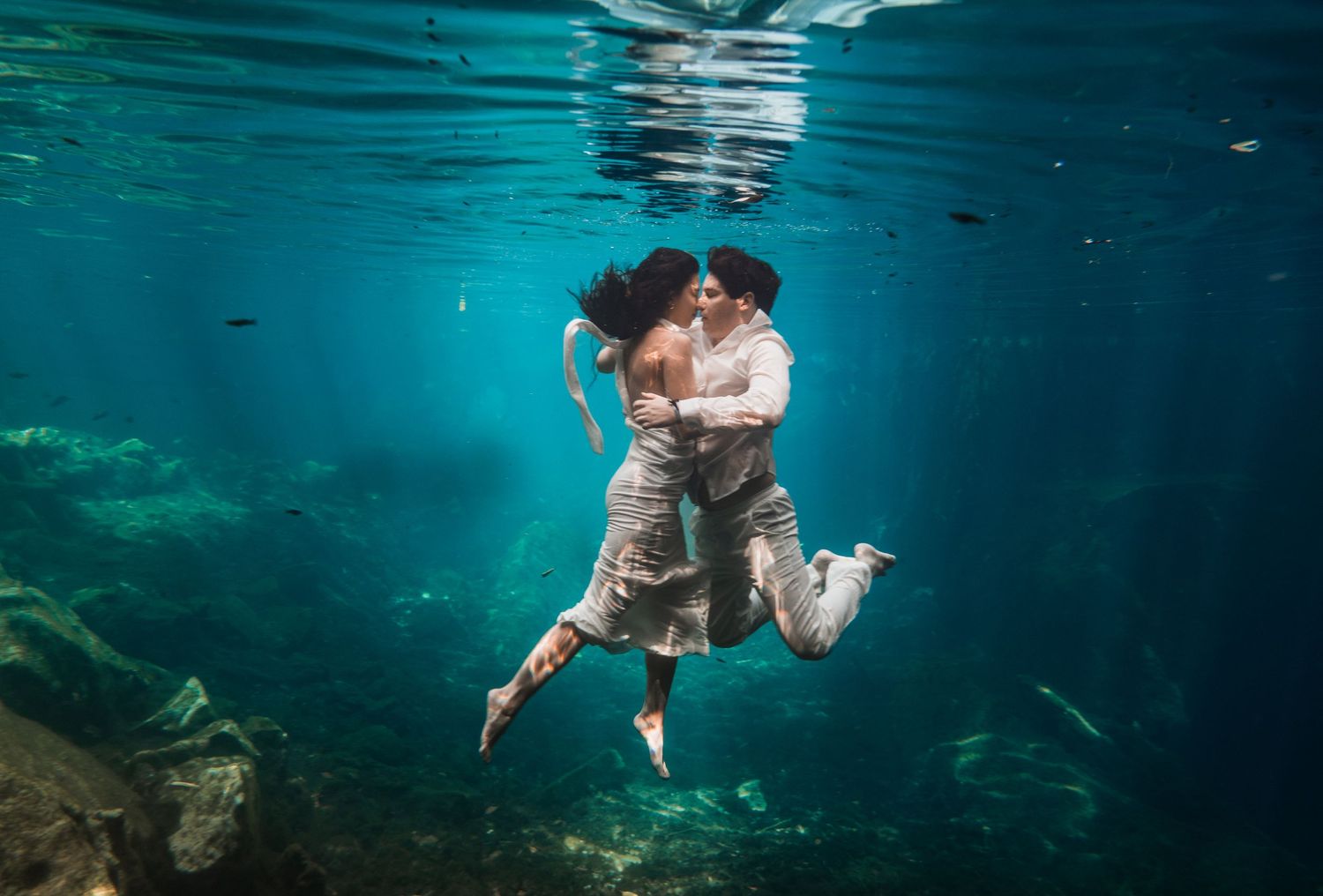 Couple embracing and kissing underwater in a clear turquoise cenote, wearing white outfits in a romantic underwater photo.