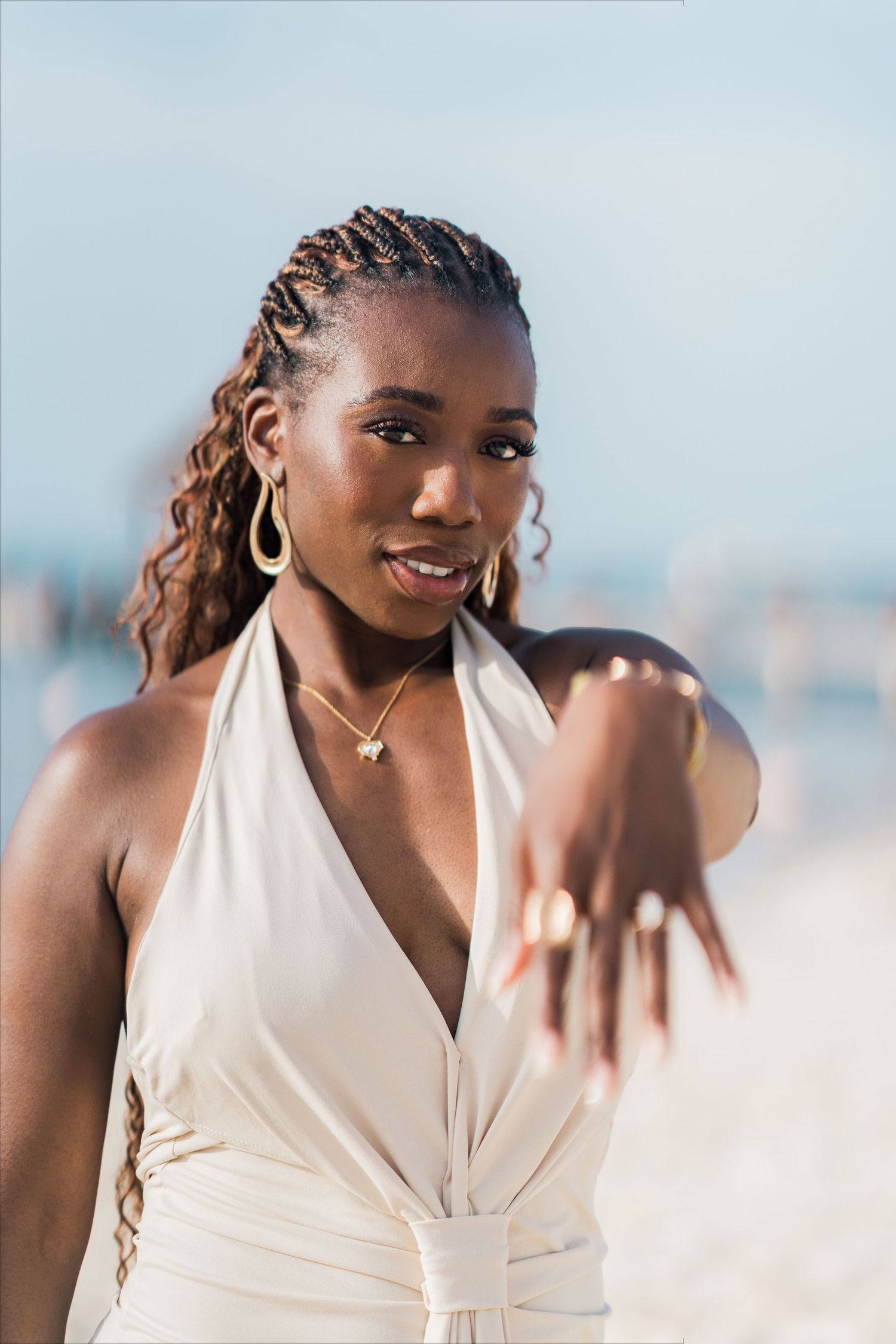 A beautiful Black woman in a white halter dress poses on a beach, showing off her engagement ring and gold accessories.