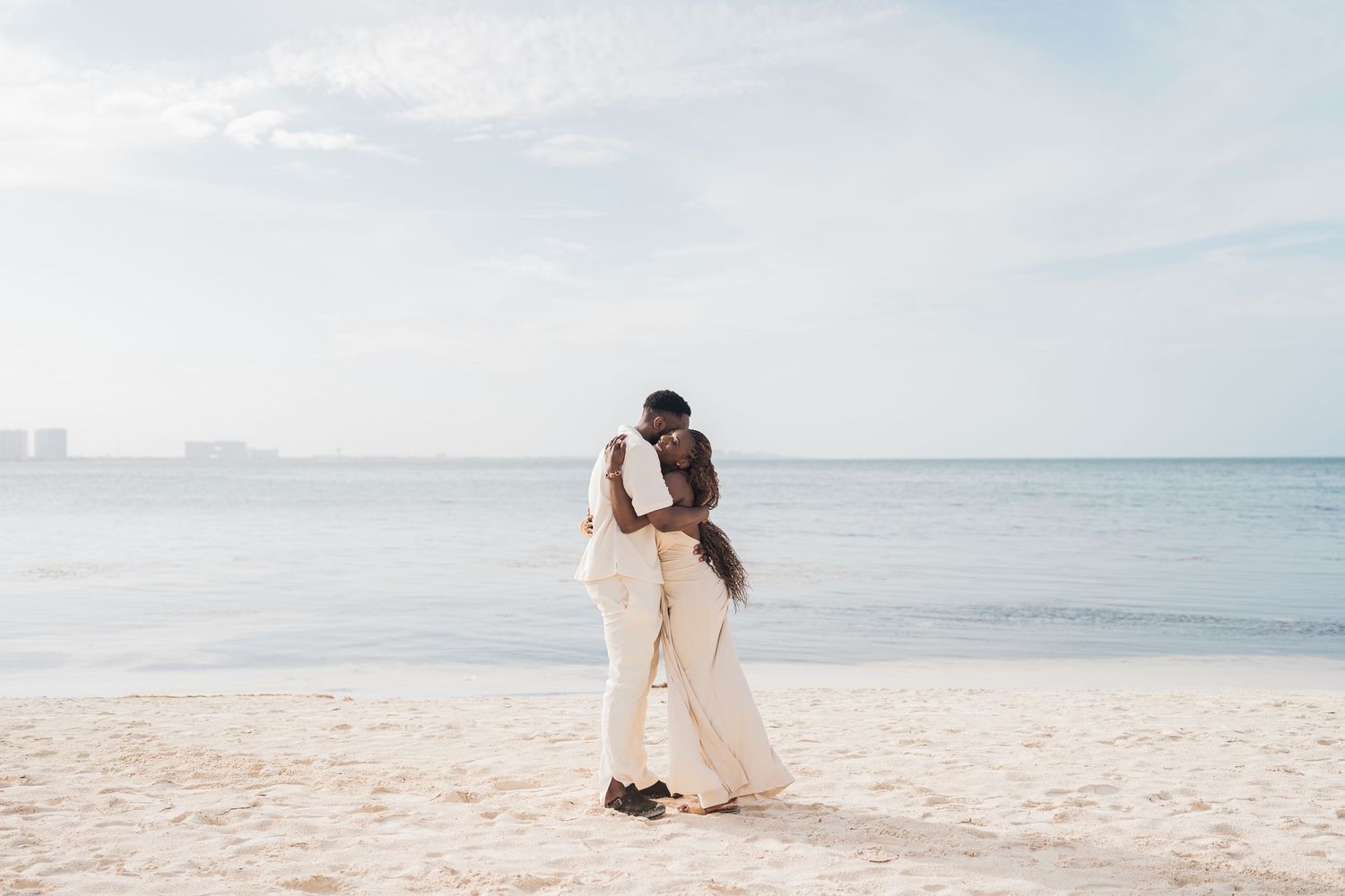 Couple sharing a romantic kiss on a sandy beach in white wedding attire with ocean backdrop.