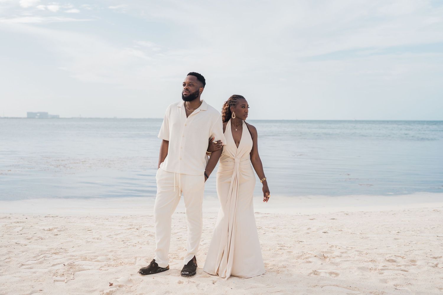 Couple in white outfits walking on a sandy beach, with calm ocean waves and a cloudy sky in the background.