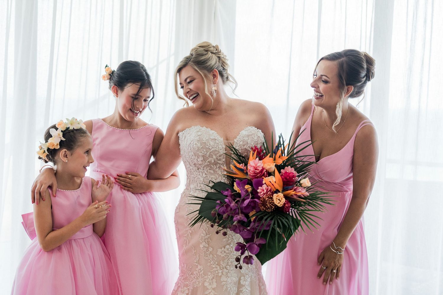 Bride in lace gown laughs with bridesmaids in pink dresses, holding tropical bouquet with roses and palm leaves.