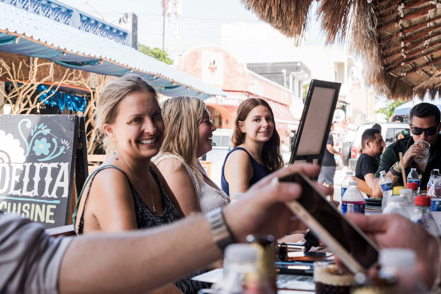 Riviera Maya Multi Day wedding - Women smiling and taking photos at an outdoor restaurant patio with tropical decor and a menu board.