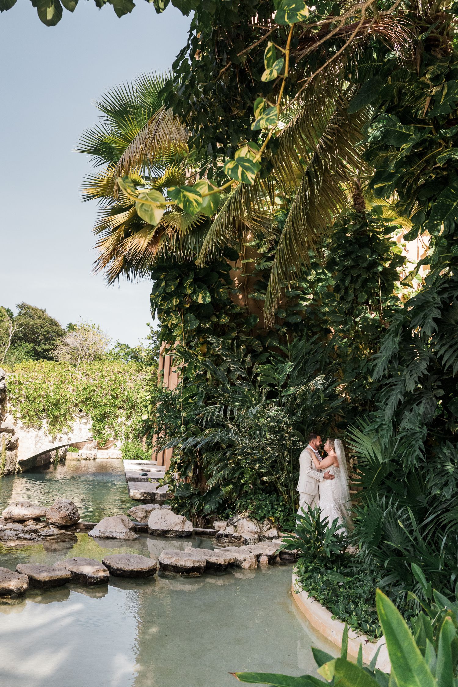 Hotel Xcaret México Wedding  - Couple embracing beside a lush tropical garden waterway with palm trees and green foliage at a resort.