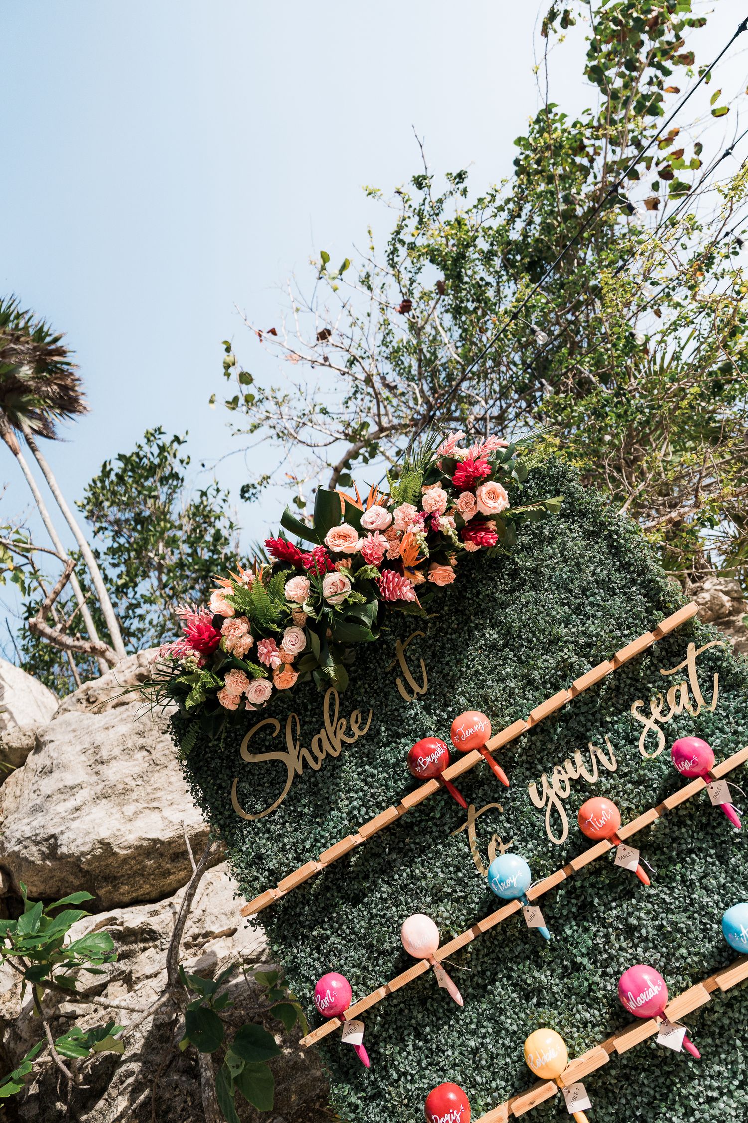 Outdoor wedding seating chart on green hedge wall with floral decor, bamboo accents, and colorful maracas under blue sky.