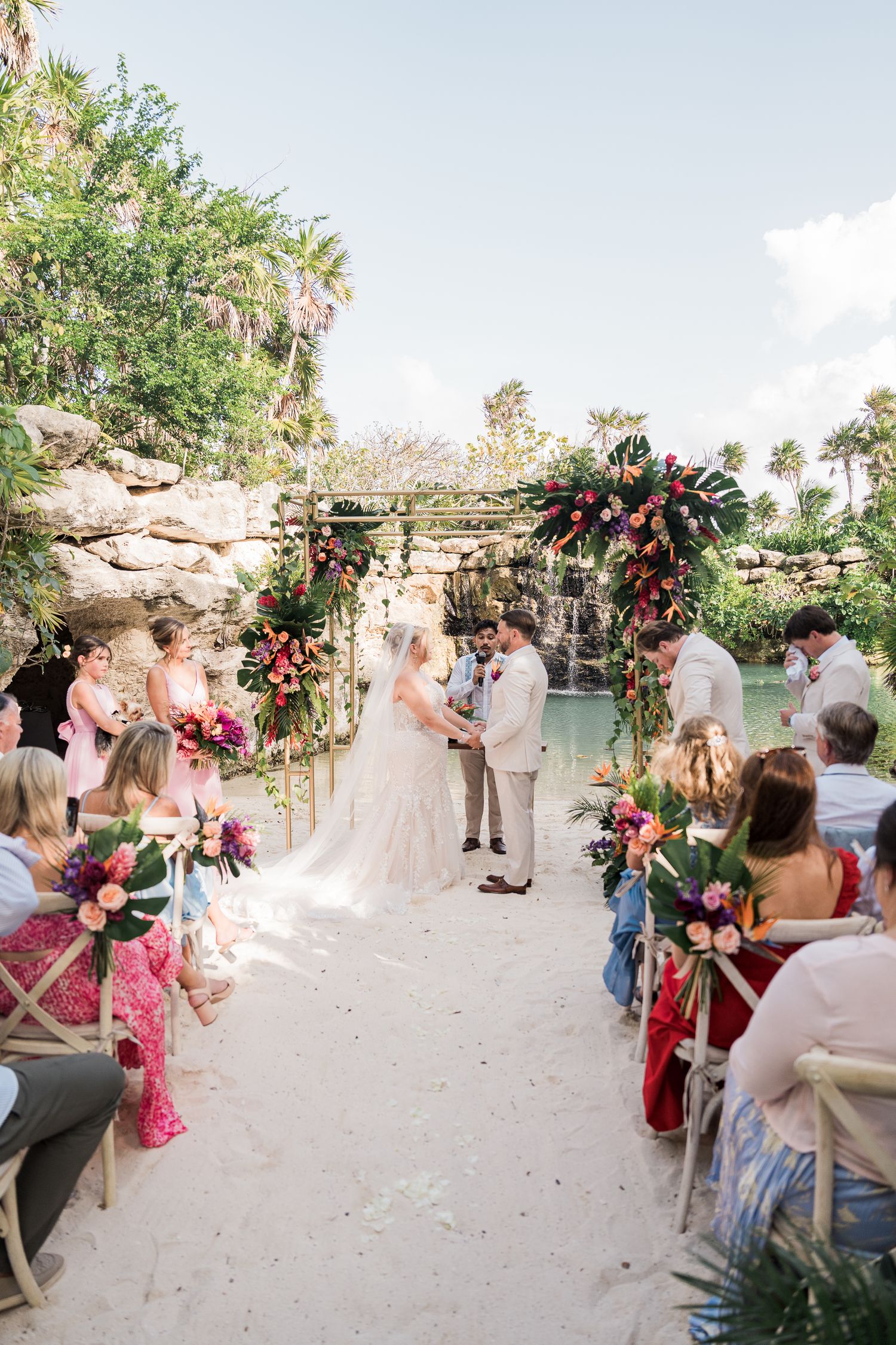 Hotel Xcaret México Wedding  - Tropical beach wedding ceremony with floral arch, waterfall backdrop, and guests seated on white sand.