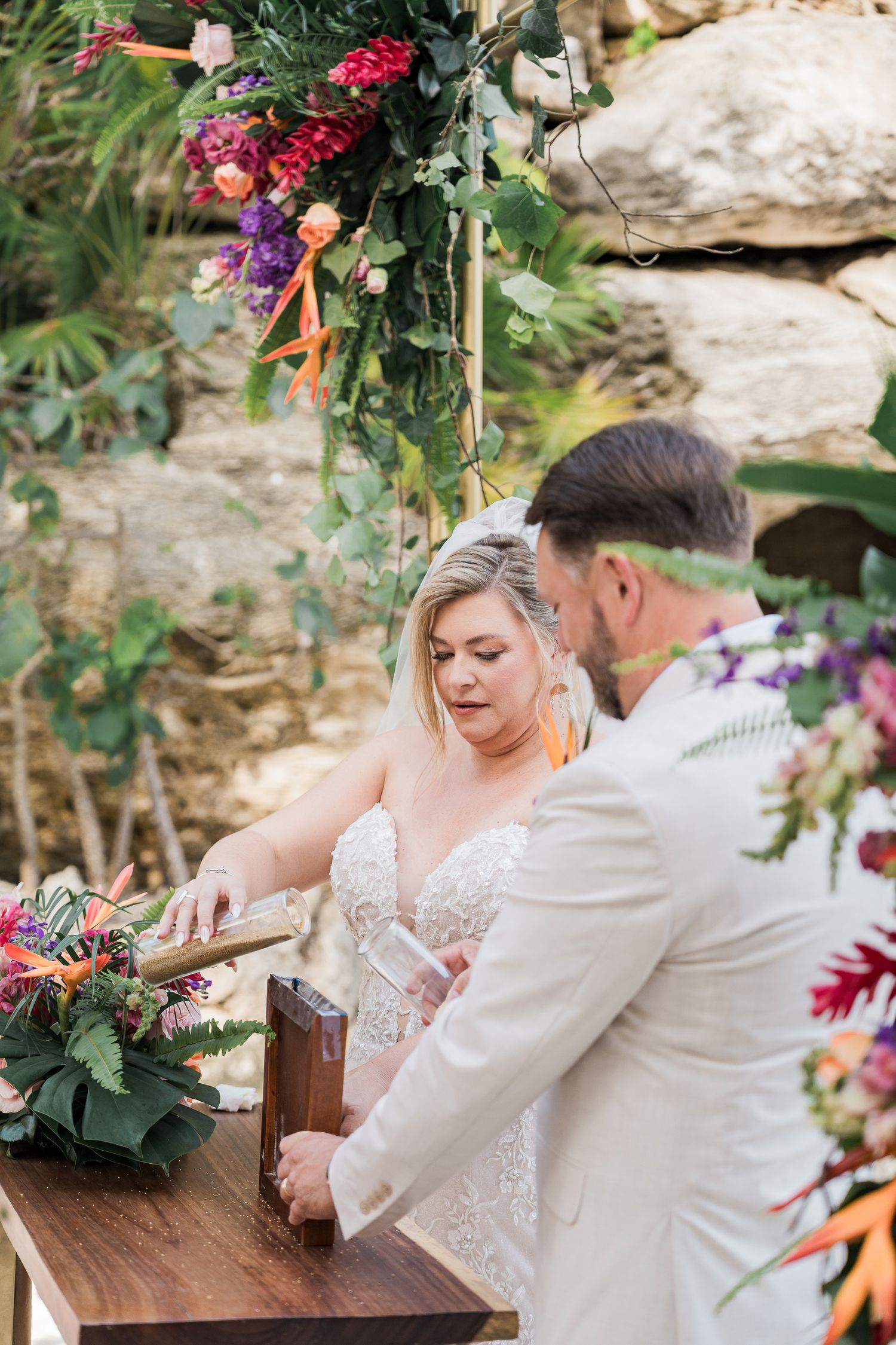 Bride and groom performing a unity sand ceremony at an outdoor wedding altar adorned with tropical floral arrangements.