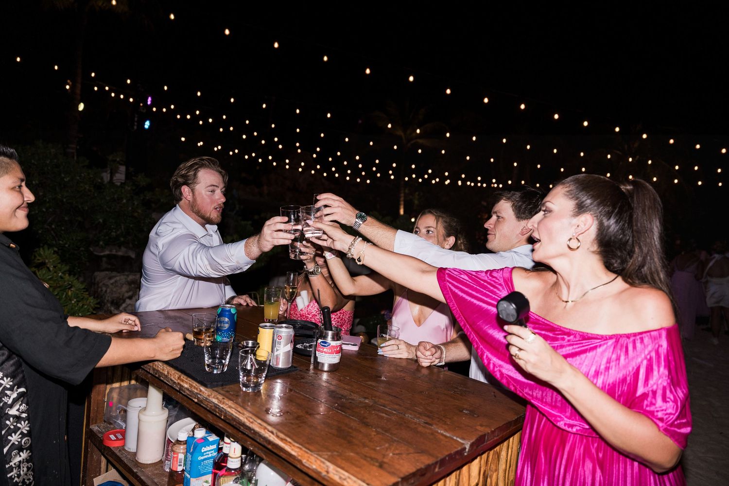 Guests in formal attire toast drinks at an outdoor wedding reception bar under string lights at night.