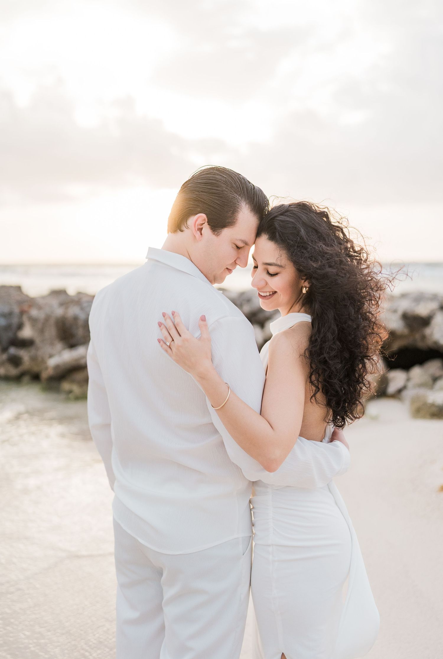 Couple in white attire embracing on a beach at sunset, smiling together near rocky shoreline.