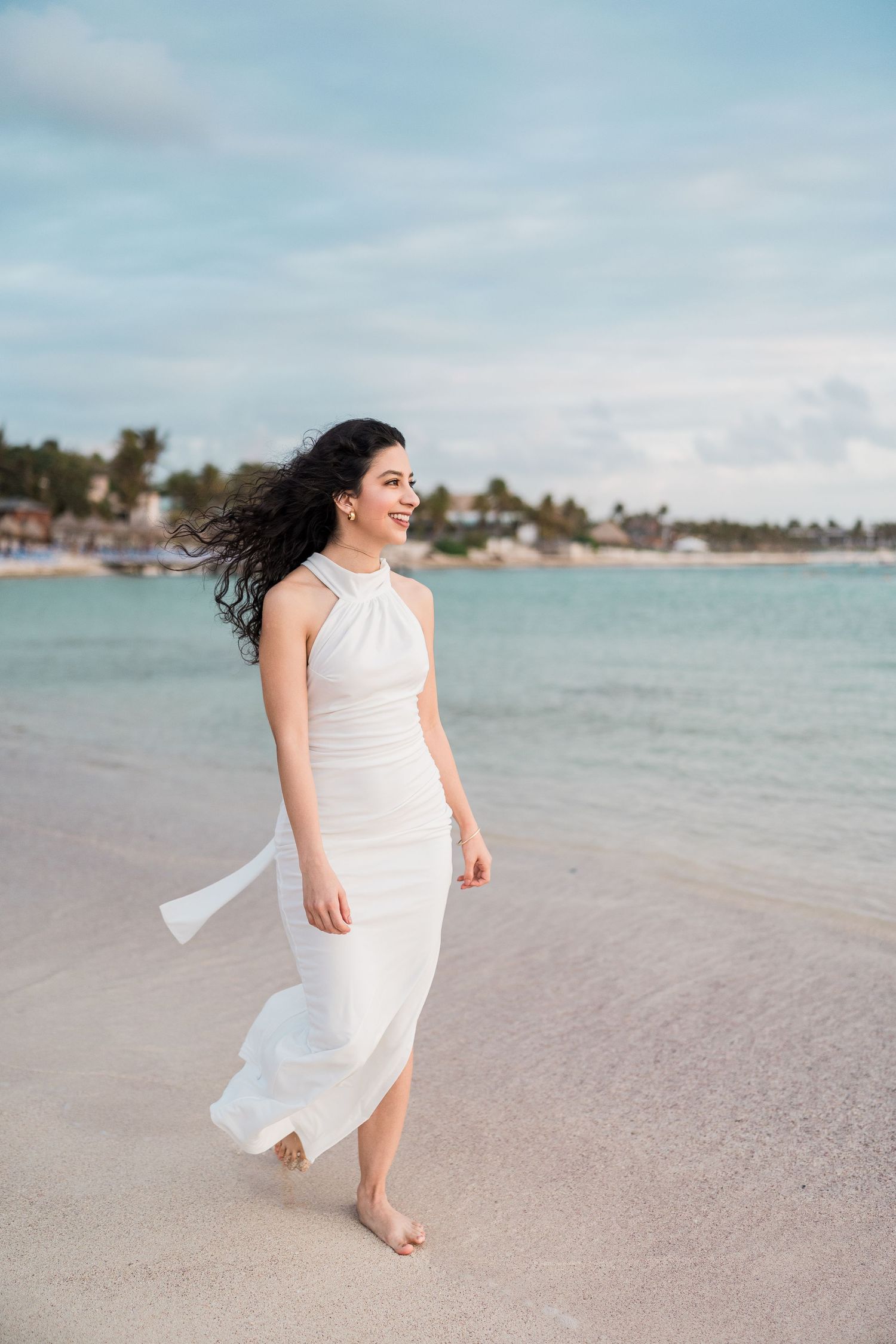 A smiling woman in a flowing white halter dress walks barefoot on a tropical beach with turquoise water.
