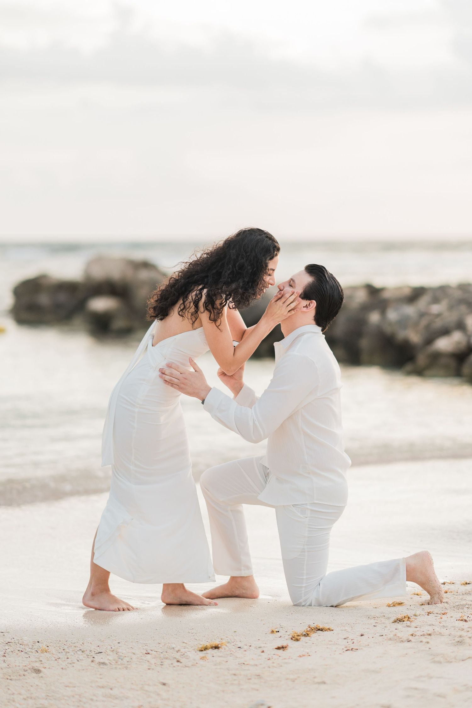 Couple in white outfits sharing a romantic beach proposal moment, man on one knee holding woman's face near rocky shore.