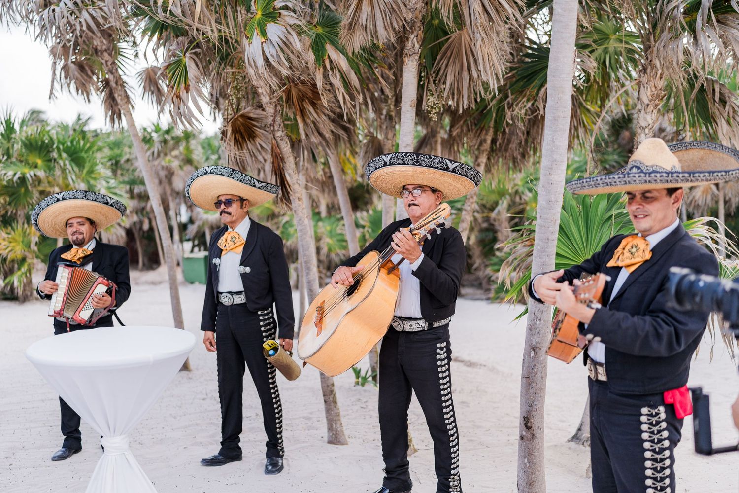 Mariachi band in traditional charro suits performing on a sandy beach surrounded by palm trees.