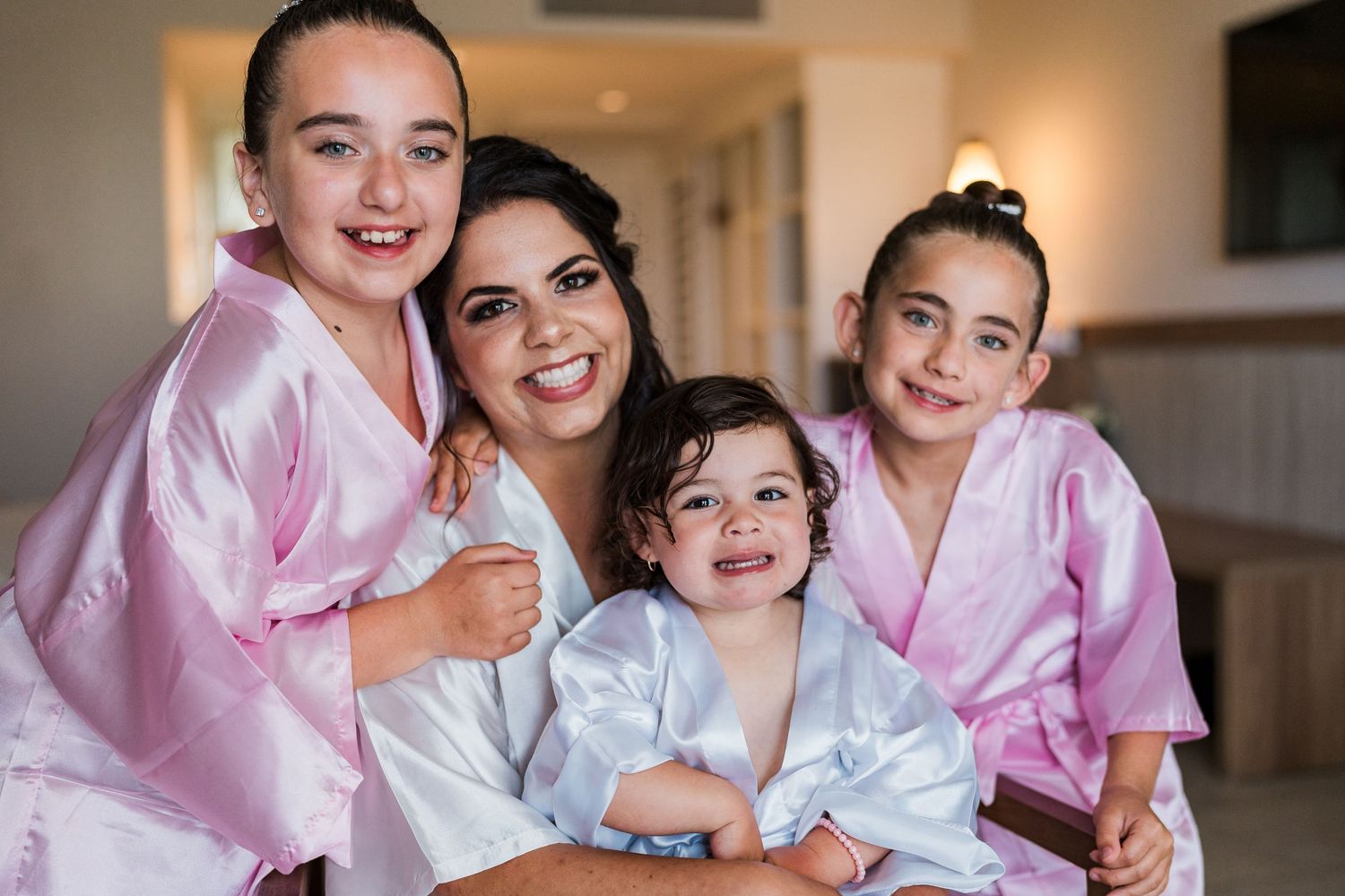 Smiling bride in white robe posing with three young girls in pink robes before wedding.