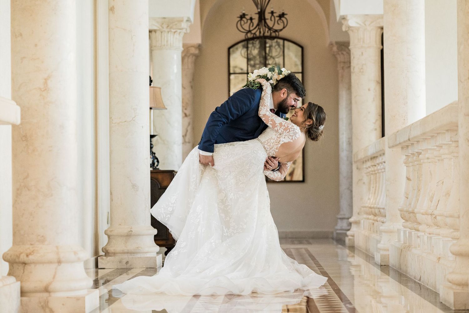 Groom dips bride for romantic kiss between marble columns in grand venue interior.