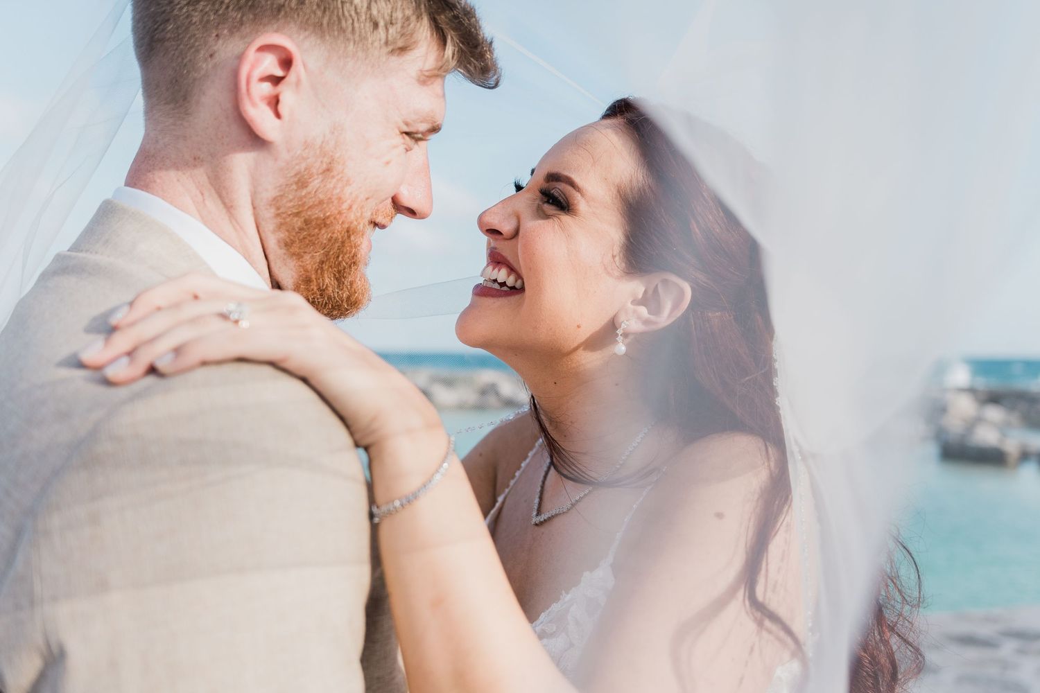 Bride and groom share a romantic laugh under the veil with turquoise ocean in the background.