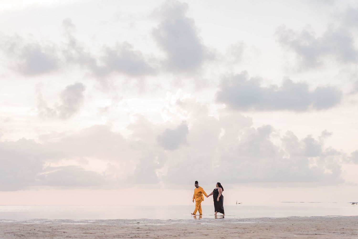 Two women holding hands walking along a calm beach at sunset.