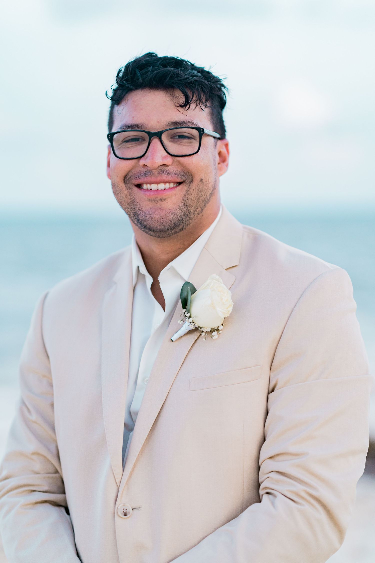 Smiling groom in beige suit with white rose boutonniere posing on beach with ocean in background.