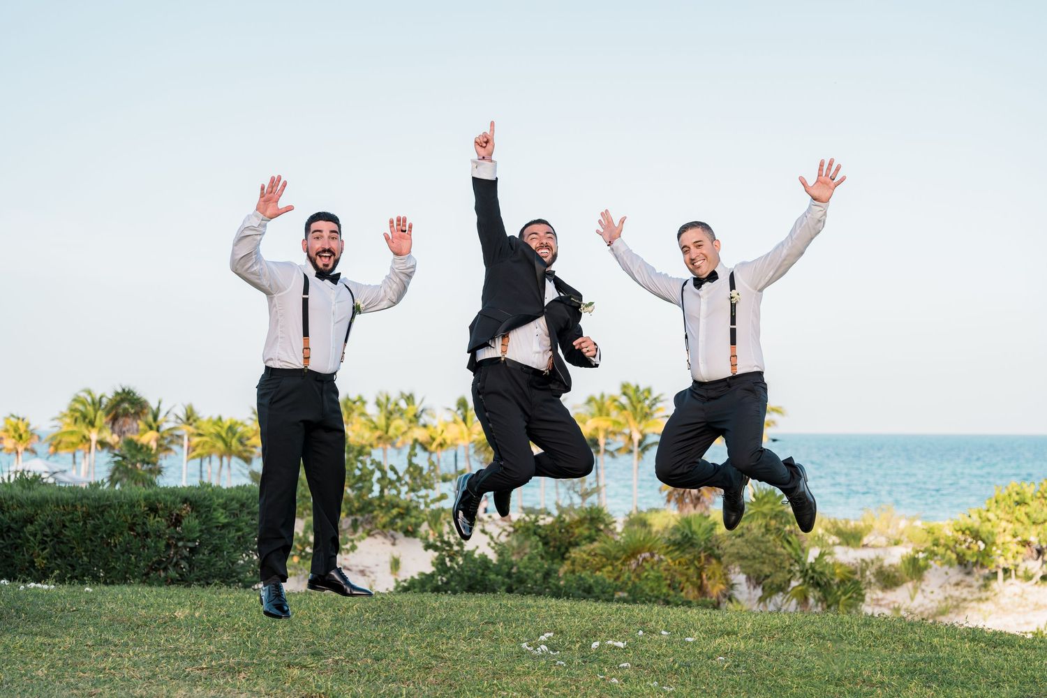Three groomsmen in black tuxedos joyfully jump in the air near a tropical beach.