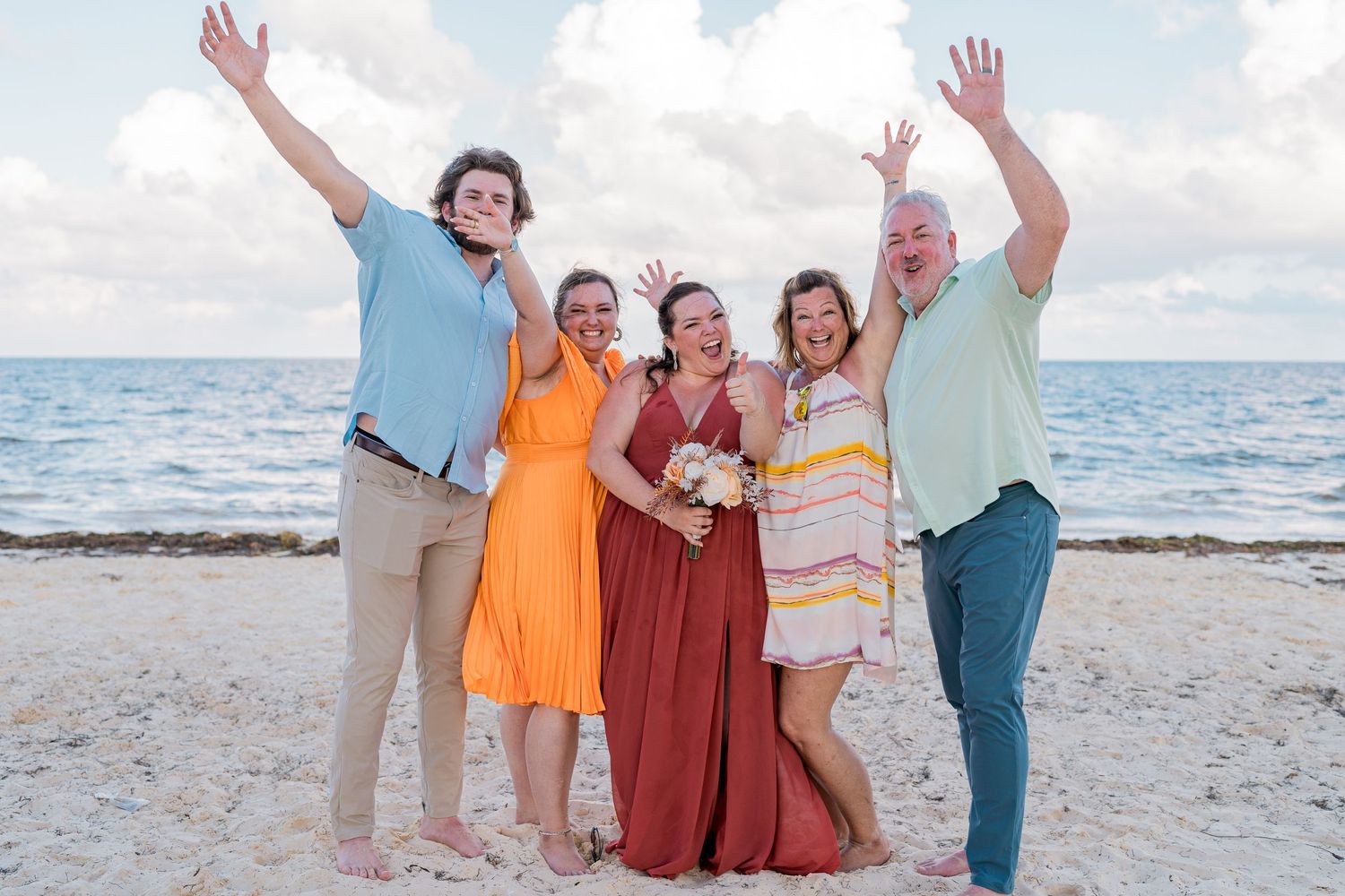 Group of five joyful wedding guests pose with raised hands on a sunny beach with ocean in background.