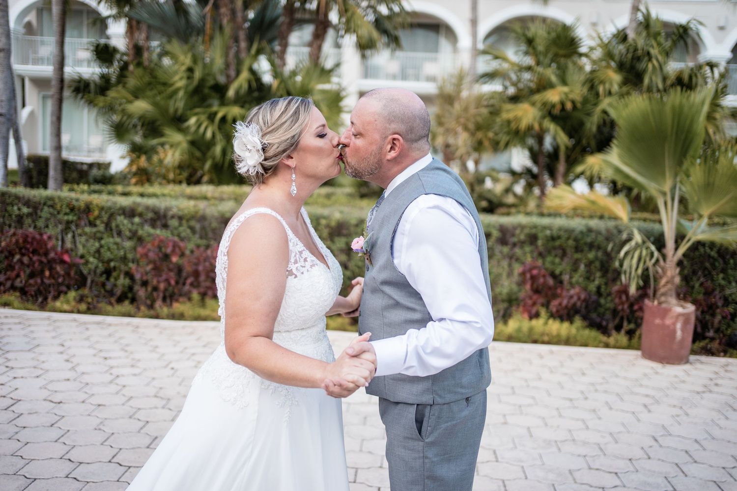 Bride and groom share romantic kiss outdoors surrounded by tropical palms and resort landscaping.