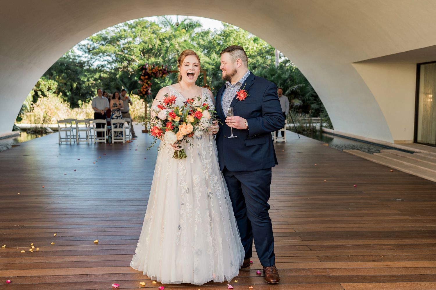 Secrets Akumal Wedding - Bride and groom laughing together under arch at outdoor wedding venue, bride holding colorful bouquet.