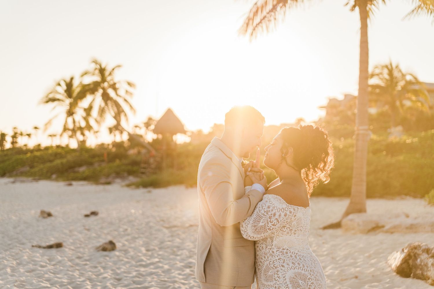 Xcaret Arte Wedding - Couple sharing a romantic kiss on a sandy beach at golden sunset with palm trees in the background.