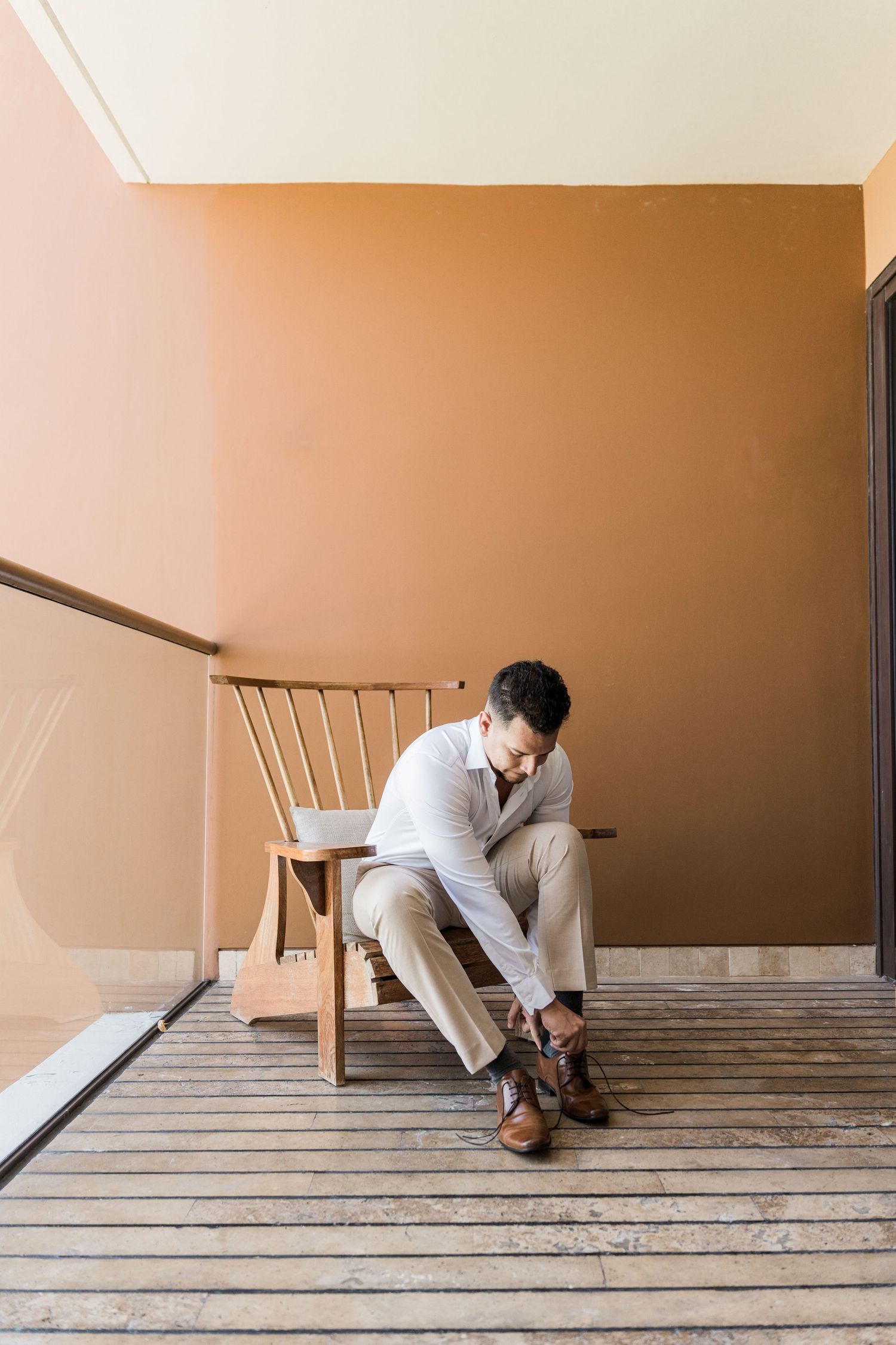Man in beige outfit sitting on wooden chair tying brown leather shoes against warm terracotta wall.