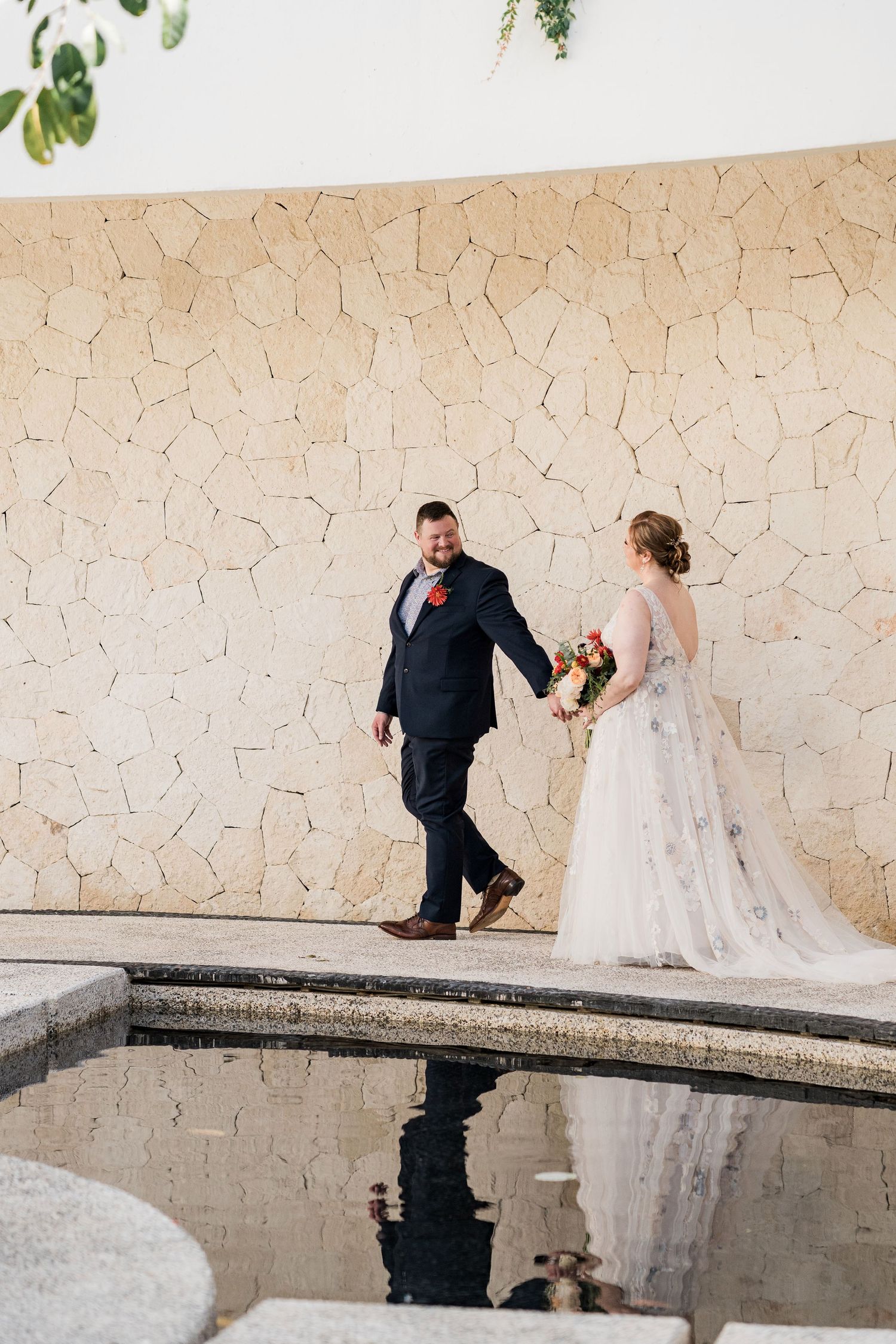 Secrets Akumal Wedding - Bride and groom walk hand-in-hand beside a reflective pool against a stone wall at their wedding venue.