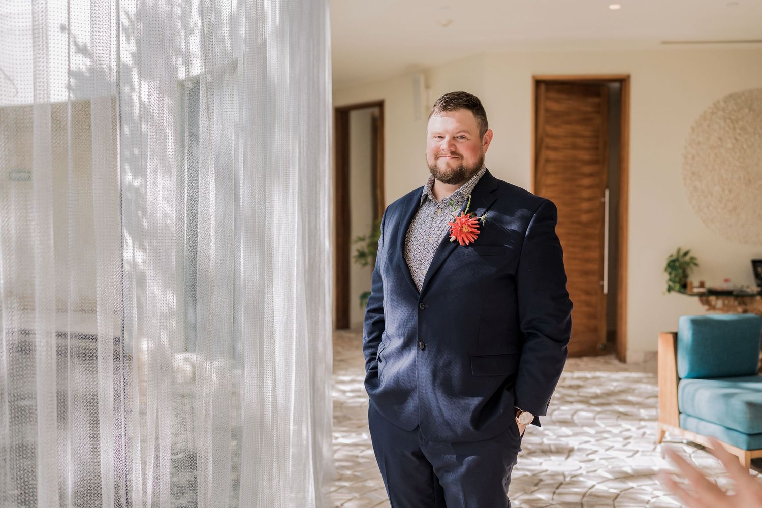 Secrets Akumal Wedding - Groom in navy suit with red boutonniere standing confidently by sheer curtains in elegant wedding venue.