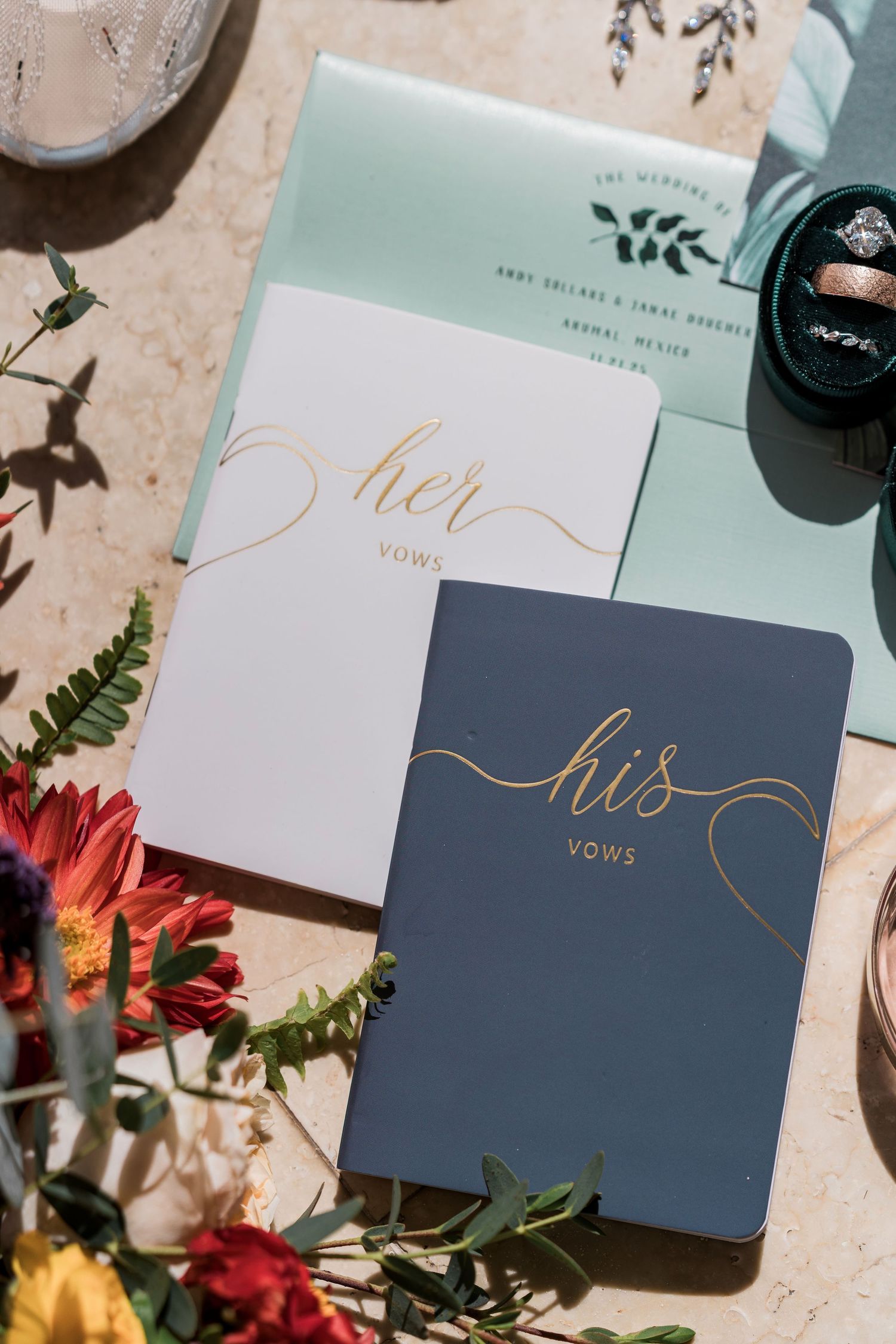 His and her gold script vow books in navy and white beside mint wedding invitations and floral arrangement.