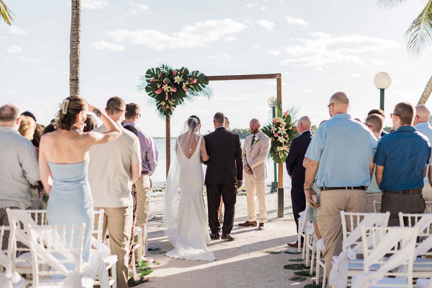 Outdoor beach wedding ceremony with guests seated in white chairs as couple exchanges vows under a floral arch.