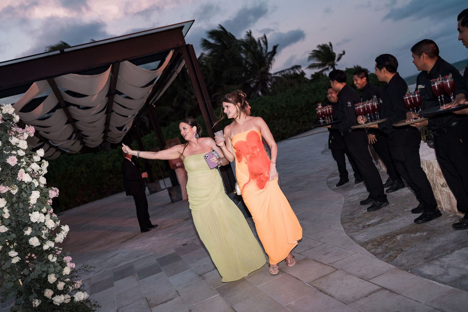 Two women in elegant gowns dance outdoors at a beachside evening event with staff holding trays nearby.