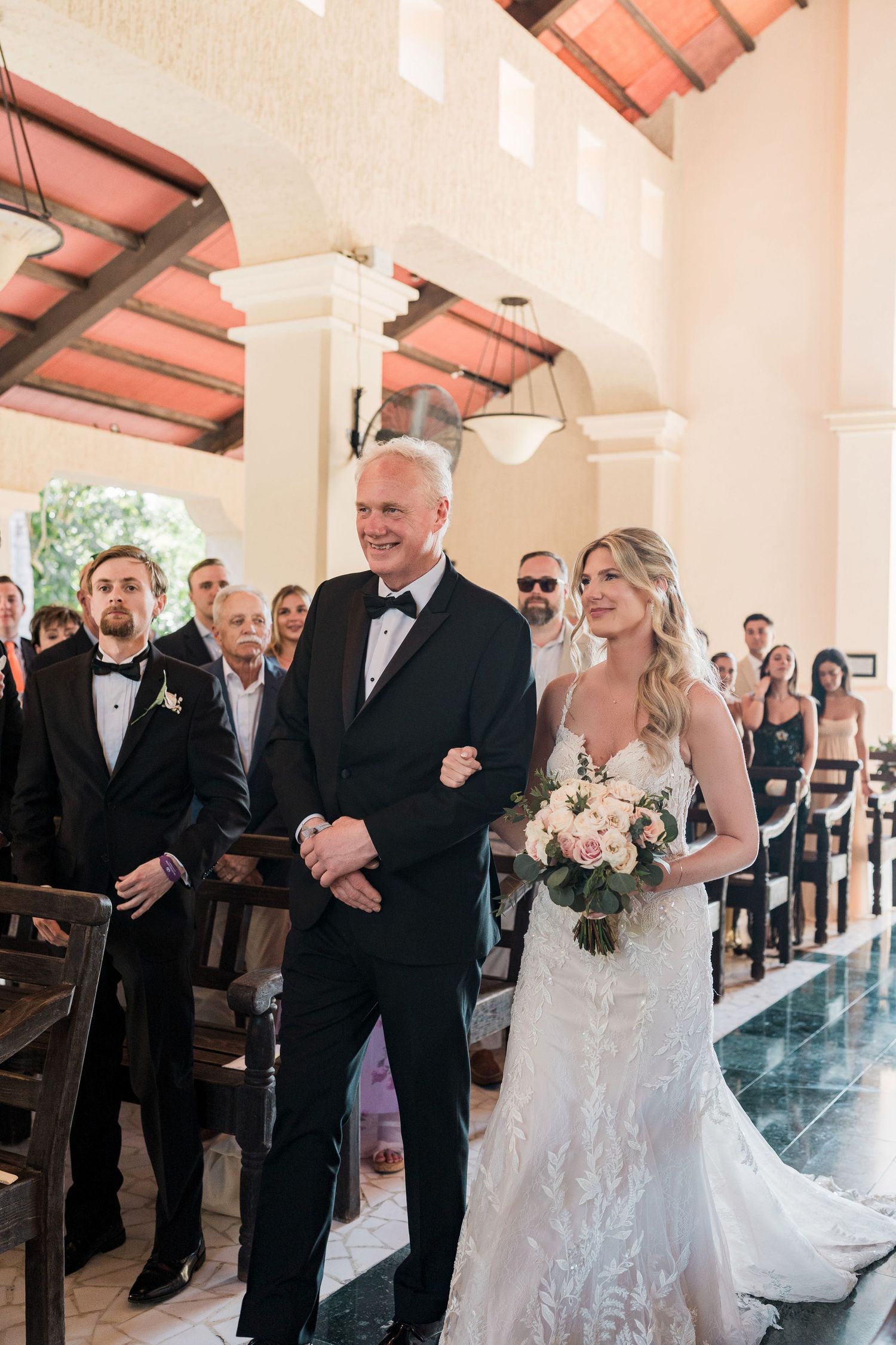 Bride in lace gown walks down church aisle with father in tuxedo, holding white bouquet at elegant wedding ceremony.