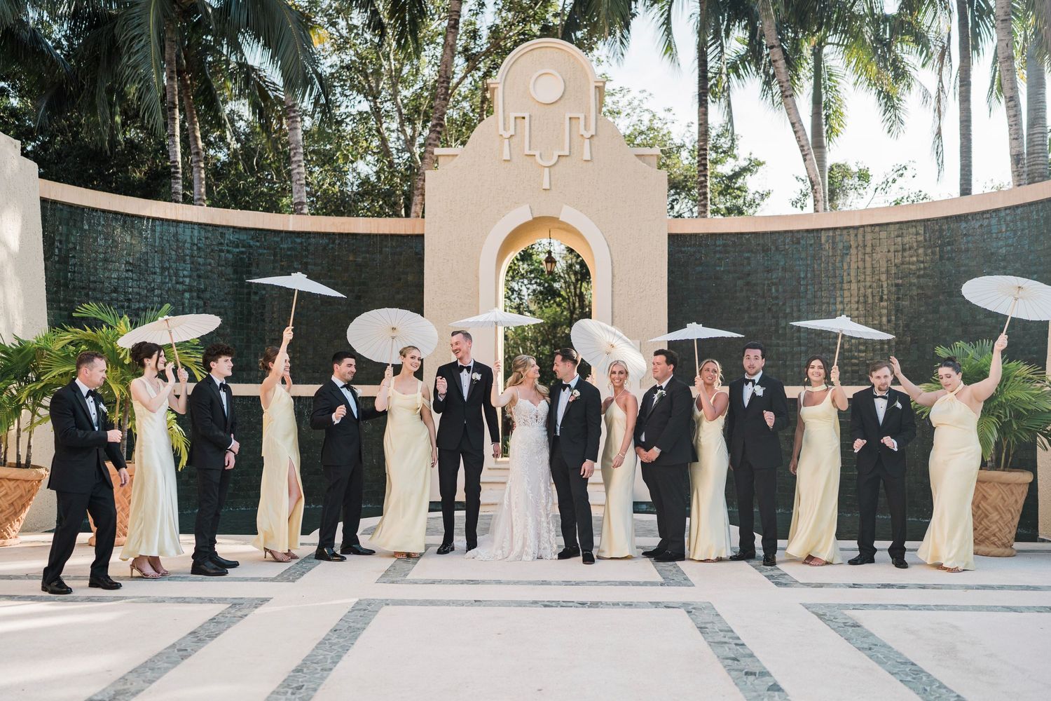 Wedding party in formal attire holding white parasols posed before elegant arch backdrop at outdoor tropical venue.