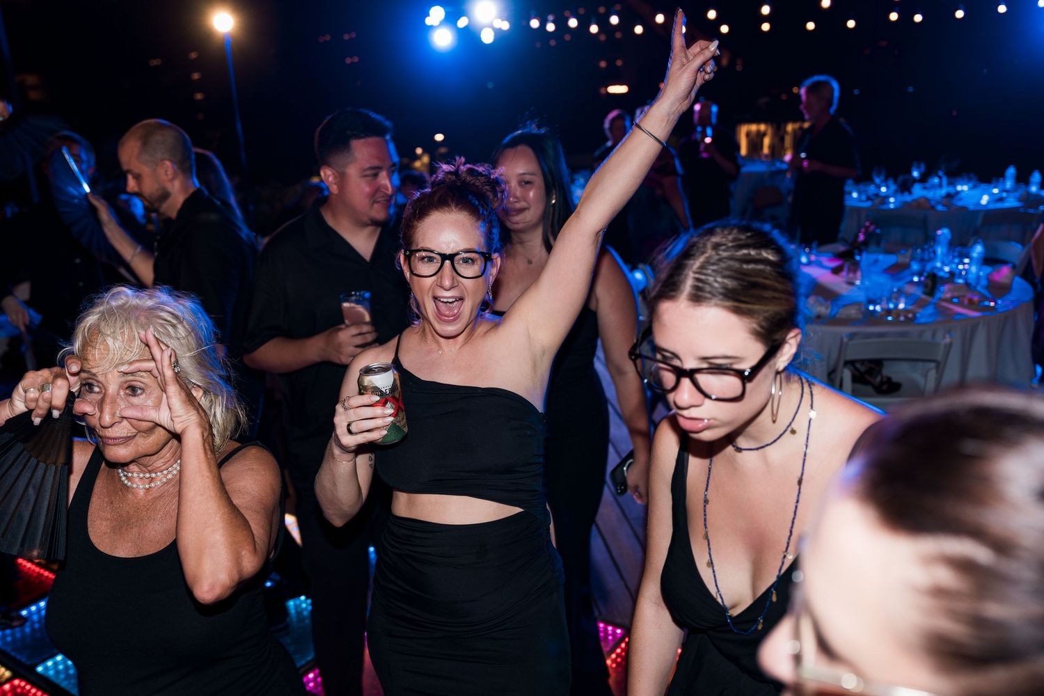 Women dancing and celebrating at a nightclub party, wearing black outfits and glasses under colorful lights.