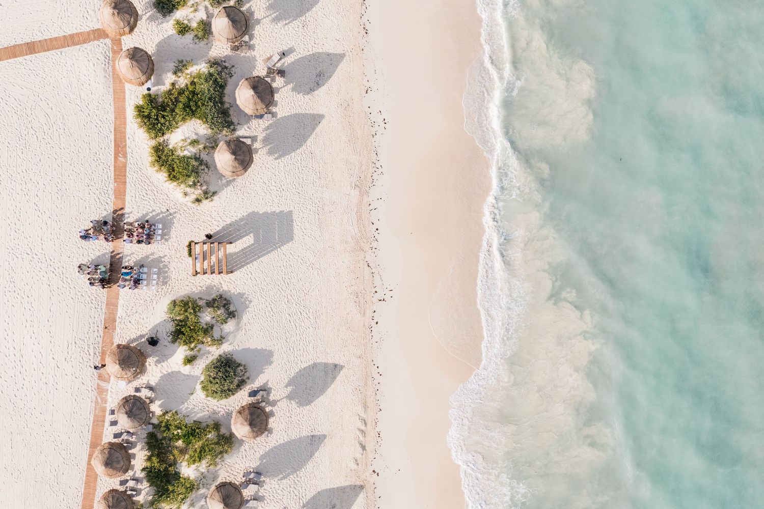 Aerial view of a pristine white sand beach with turquoise waves, palm umbrellas, and lounge chairs casting long shadows.