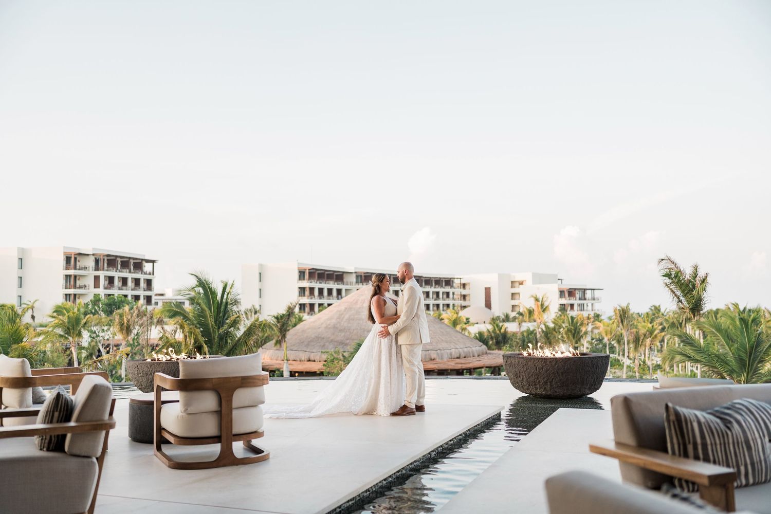 Bride and groom share romantic moment on luxury resort terrace with pool, palm trees, and ocean view backdrop.
