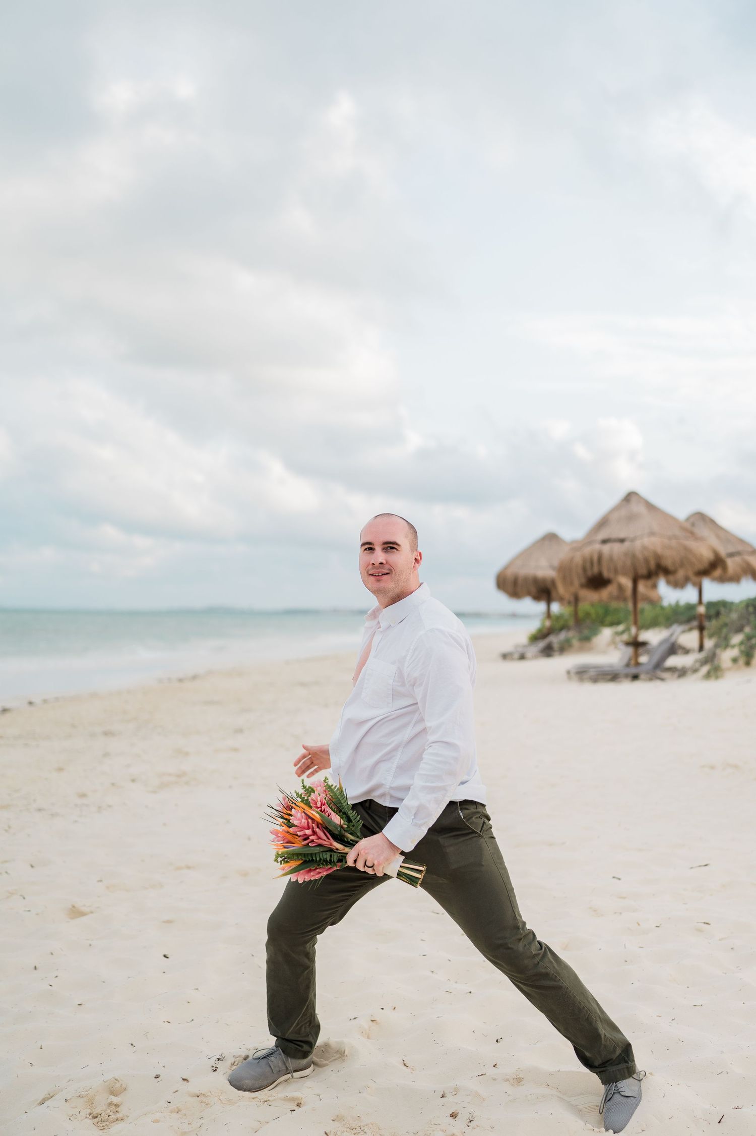 Groom in white shirt and green pants holds tropical bouquet on sandy beach with thatched umbrellas.