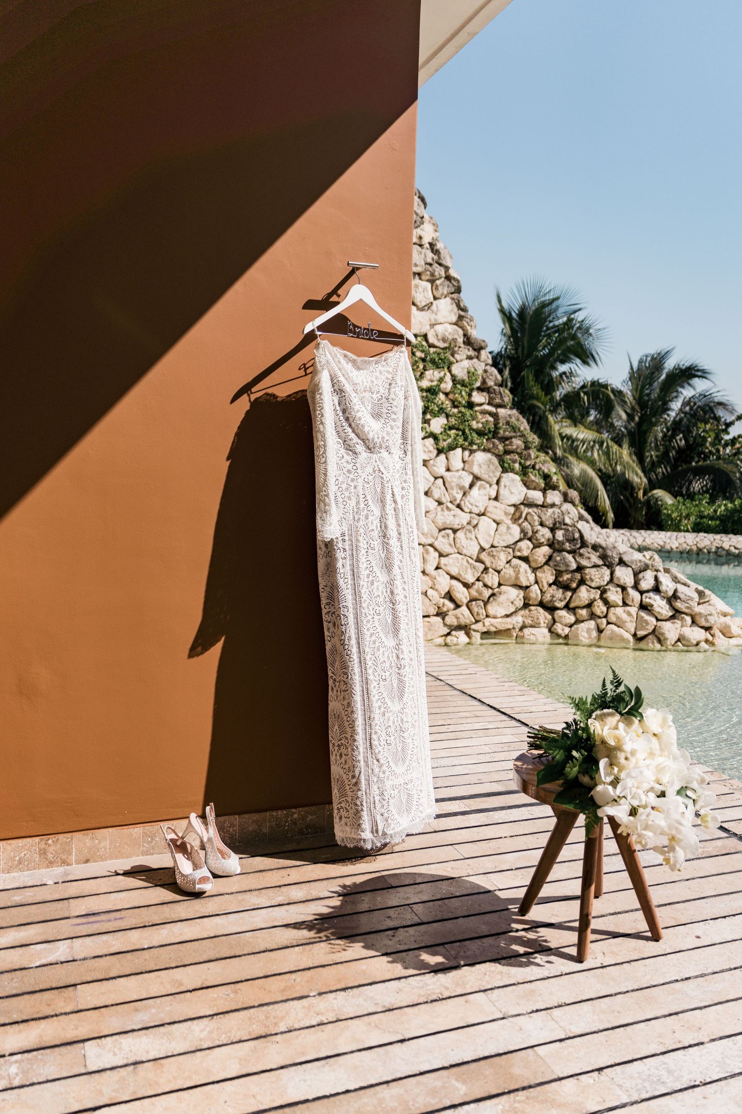 White lace wedding dress hanging outdoors at Xcaret Mexico resort with bridal bouquet and heels on wooden deck.
