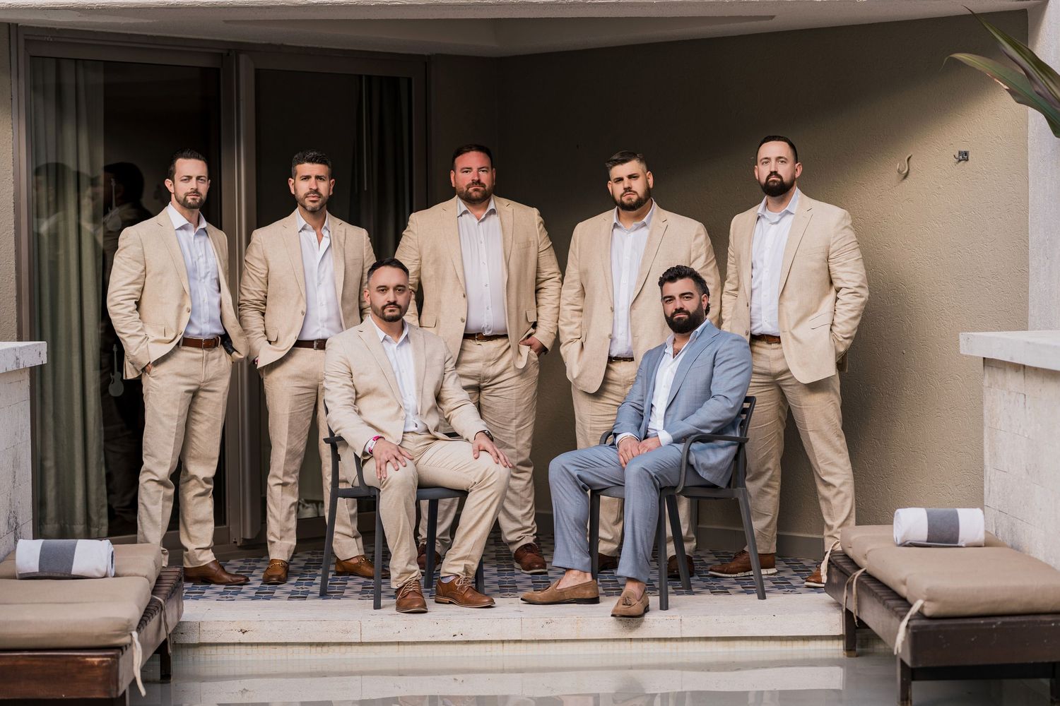 Group of groomsmen in tan suits posing on a balcony, two seated in front, five standing behind. Hard Rock Riviera Maya wedding photographer