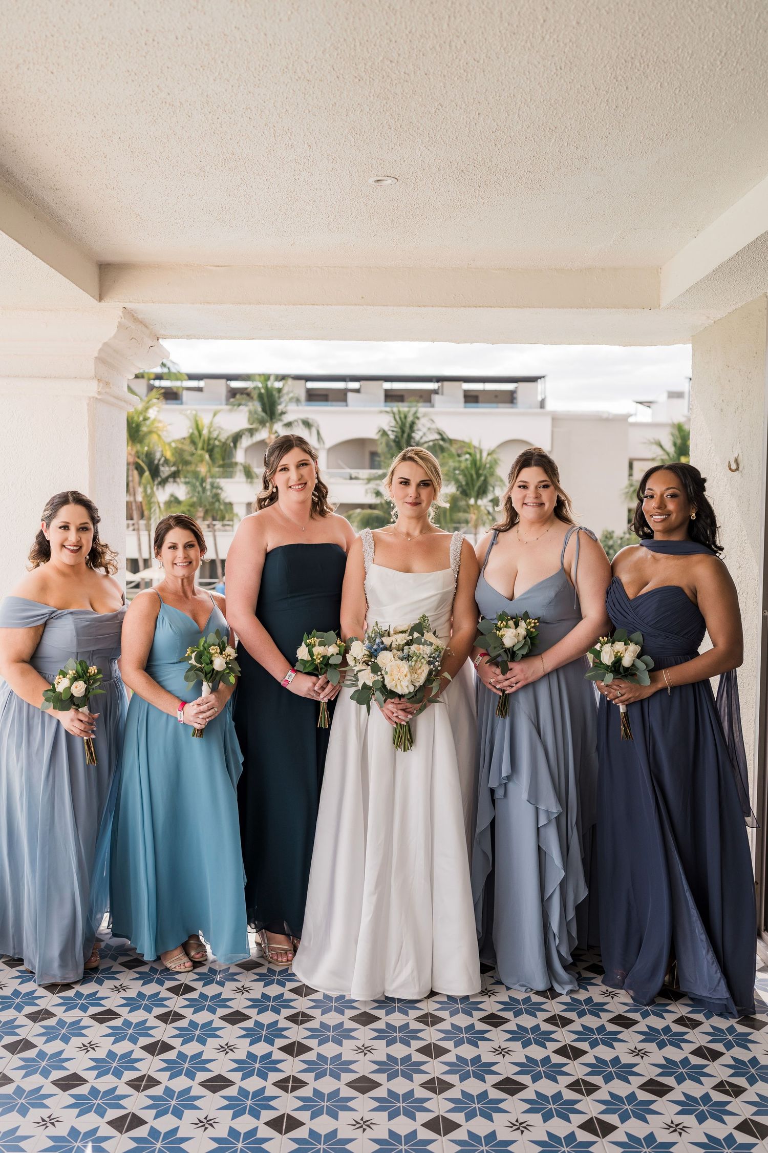 Bride in white gown poses with five bridesmaids in blue dresses holding bouquets at a tropical wedding venue.