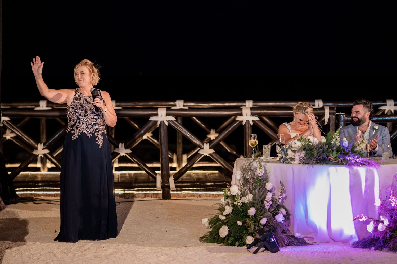 Woman giving a speech at an outdoor wedding reception at night, with the bride and groom seated at a decorated table.
