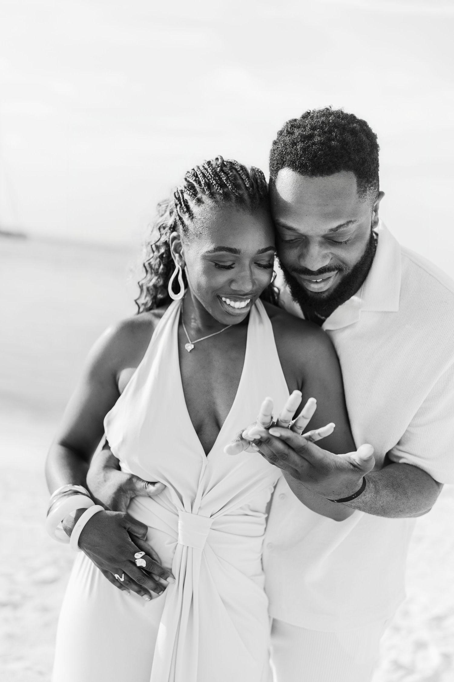 Black and white photo of a happy couple on the beach, man embracing woman from behind as they look at an engagement ring.