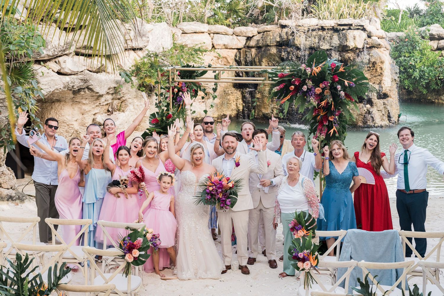 Hotel Xcaret México wedding - Joyful wedding party celebrating outdoors at a tropical venue with colorful florals, and a waterfall backdrop.