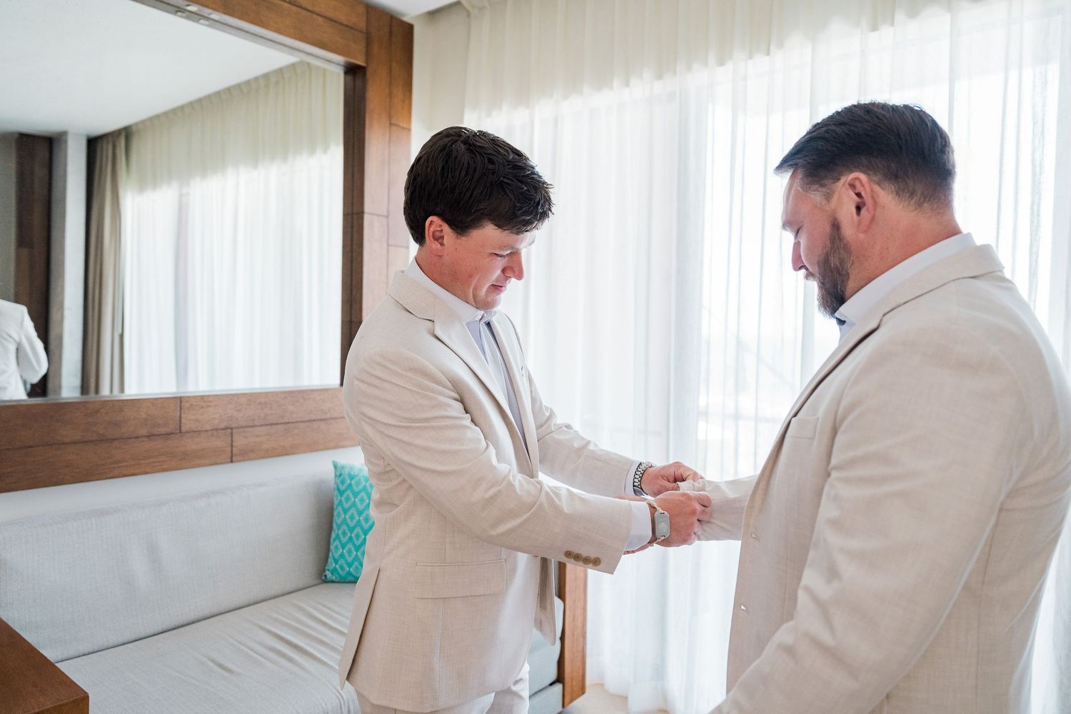 Hotel Xcaret México Wedding  - Two men in matching beige suits shake hands in a bright room, preparing for a wedding ceremony.